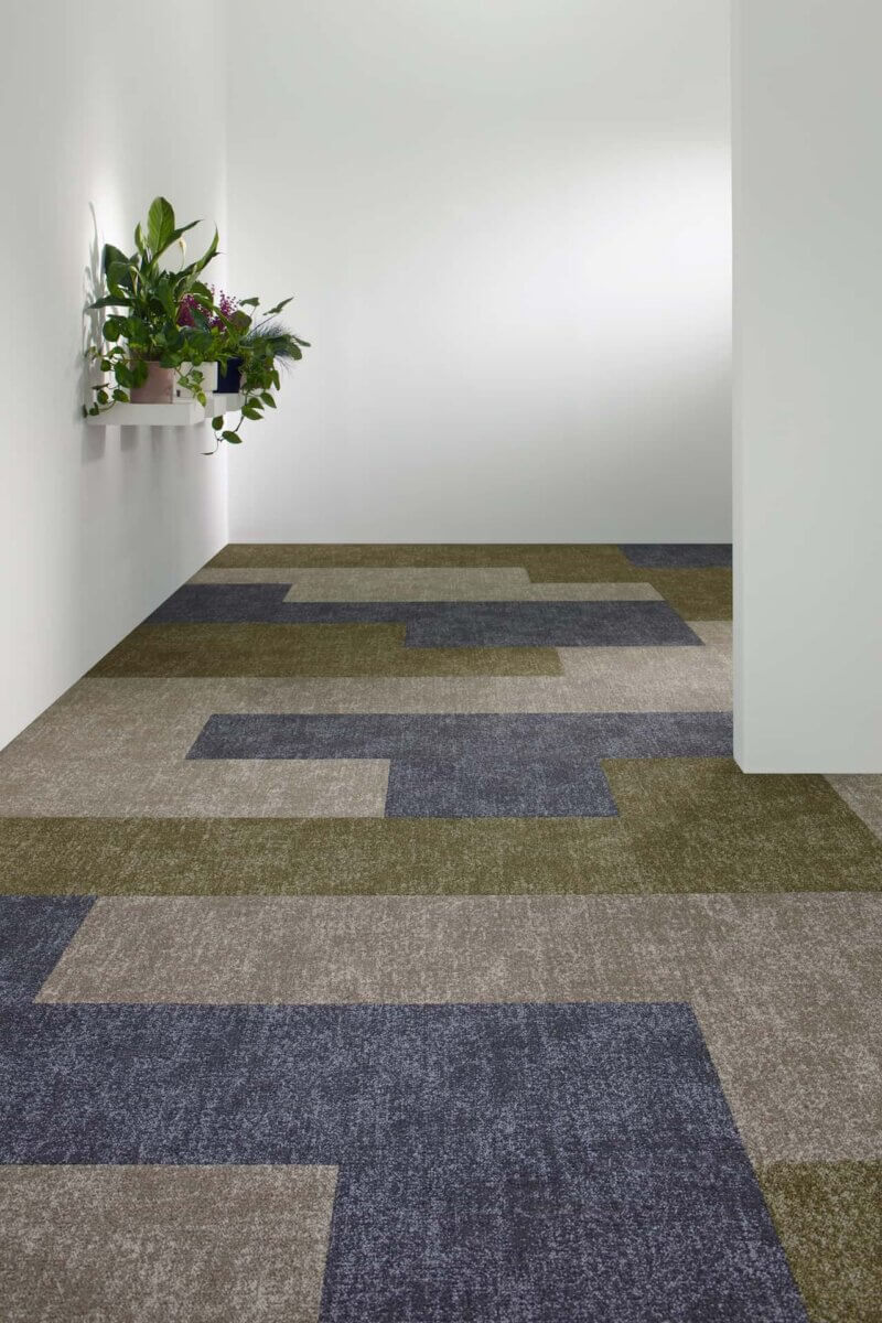 Contemporary interior hallway featuring Milliken patterned carpet tiles in layered neutral and blue tones, creating a geometric floor design, with clean white walls and planted shelving.