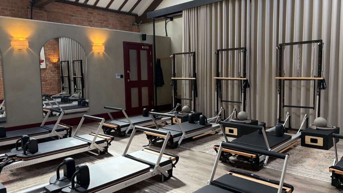 Interior of the reformer studio at Feal Pilates in Moseley, Birmingham, with rows of reformer beds and wall-mounted tower units on the Plus Floor Formation plank LVT in the silvery clay tone, framed by an arched mirror, pleated curtains, wall lights and a deep red door.