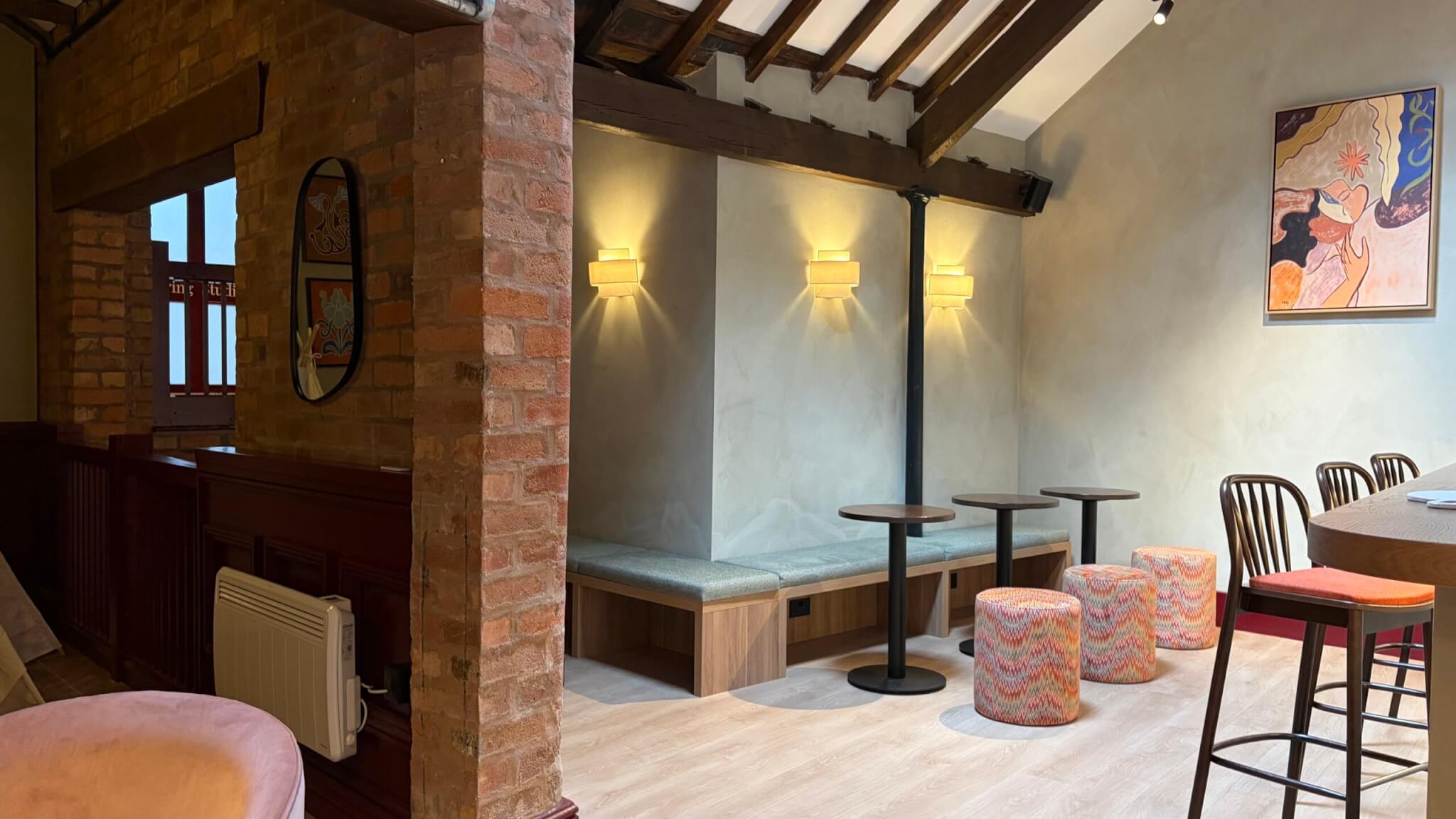 View of the wellness bar seating area at Feal Pilates in Moseley, Birmingham, with the Plus Floor Formation plank LVT in the soft pastel tone under built-in banquette seating, round tables, patterned stools and bar chairs, framed by an exposed brick pillar and wall lights beneath timber beams.